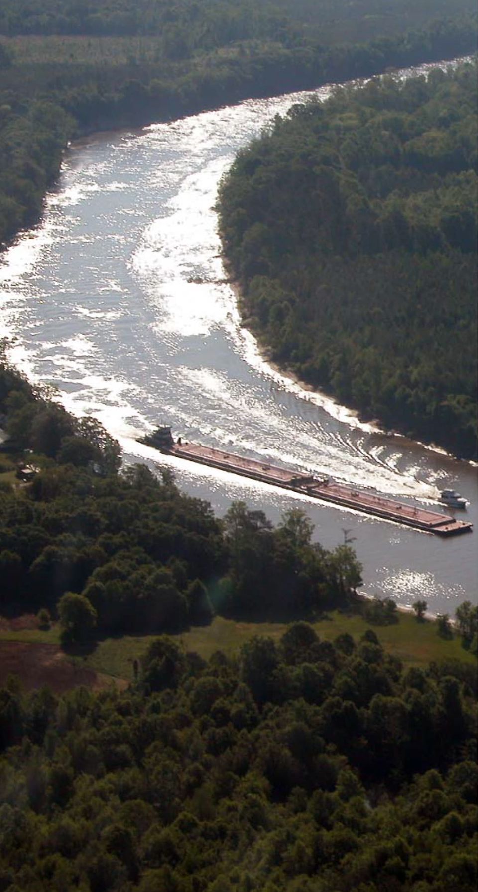 A loaded tow rounds a sweeping, sunlit bend of the waterway between wooded banks.