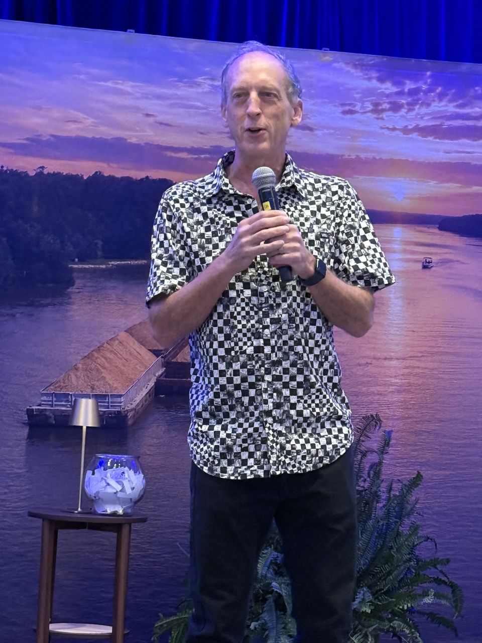 A speaker holds a microphone in front of the river backdrop.