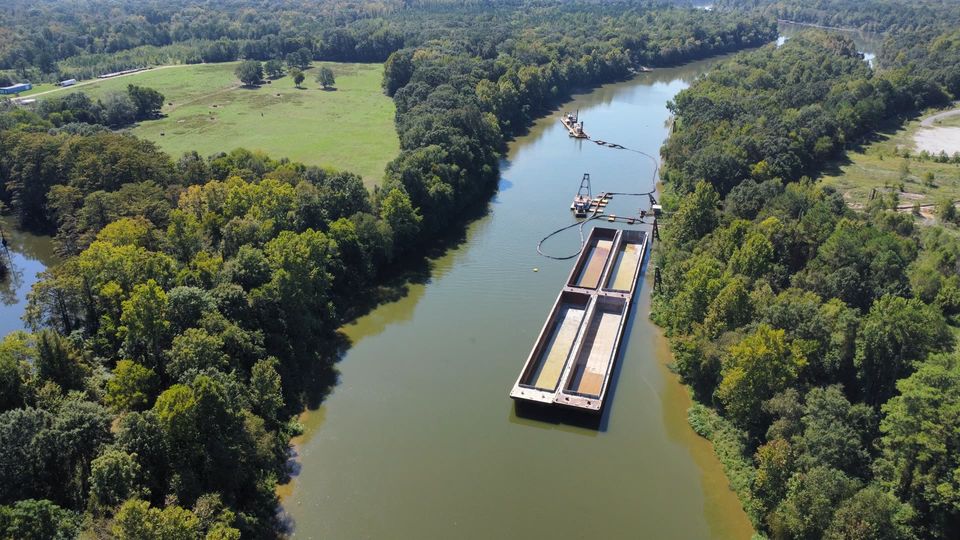 Aerial view of the waterway beside open land and a barge loading area, with forest and farmland beyond.
