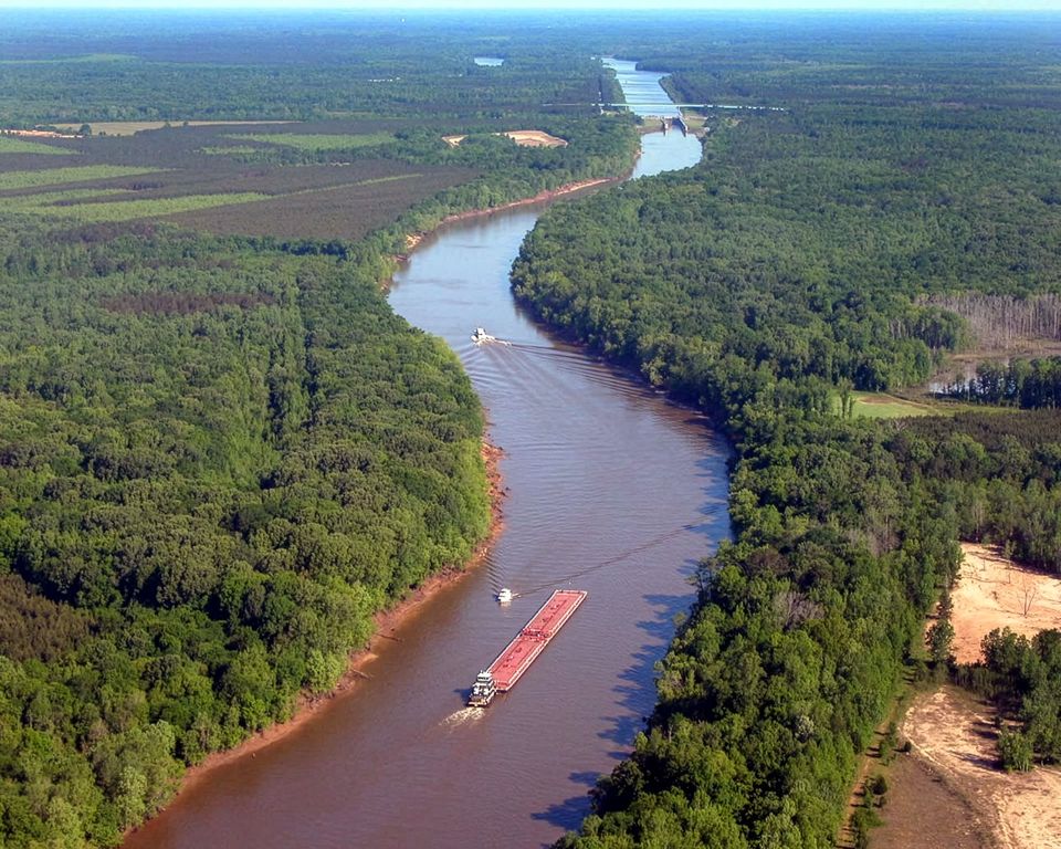 A towboat pushes a line of barges through a forested, red-banked bend of the Tennessee-Tombigbee Waterway.